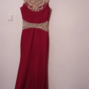 Vinröd långklänning med spetsdetaljer - Elegant vinröd långklänning med vackra spetsdetaljer i guld. Klänningen har en figurnära passform och är ärmlös med en öppen rygg. Perfekt för festliga tillfällen som nyår eller bal. Materialet är mjukt och följsamt, vilket ger en lyxig känsla.