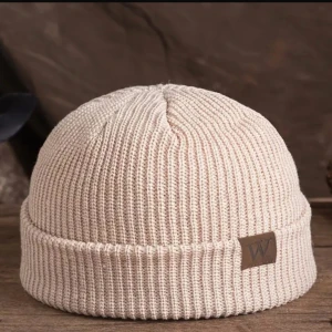 Beige ribbad mössa med logotyp - Snygg beige ribbad mössa med en stilren läderdetalj som har en 'W' logotyp. Perfekt för att hålla värmen under kyliga dagar. Den har en klassisk design som passar till det mesta i garderoben.