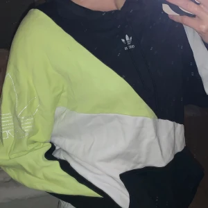 Svart/vit och neongul hoodie från Adidas - Cool hoodie från Adidas i svart/vit och neongul. Har en stor logga på ärmen och klassisk Adidas-logga på bröstet, har även luva samt ”vanlig” hoodie ficka fram.