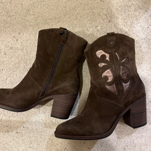 Bruna boots med broderad detalj - Snygga bruna cowboy boots i mocka med en dekorativ broderad detalj på sidan. De har en bekväm klack och dragkedja för enkel på- och avtagning. Perfekta för en stilren look! Helt nya! Säljer pga har så många boots redan
