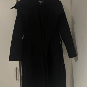 Svart kappa från ONLY - Snygg svart kappa från ONLY i mycket bra skick. Perfekt för höst och vinter med sin eleganta och tidlösa design. Den har långa ärmar och en stilren passform som passar både till vardags och fest. Ett måste i garderoben för den stilmedvetna!