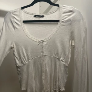 Vit långärmad topp från Ginatricot - Säljer en söt vit långärmad topp från Ginatricot med spetsdetaljer och en liten rosett framtill.  Toppen har en v-ringad design och är gjord i ett mjukt material. 