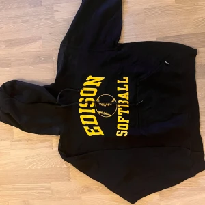 Rusell - Svart hoodie med gul Edison Softball-tryck - Säljer en svart hoodie från Gildan med ett gult Edison Softball-tryck på framsidan. Den har en klassisk känguruficka och justerbar huva med dragsko. Perfekt för en avslappnad stil.