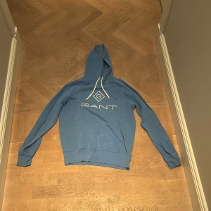 Blå hoodie från GANT - Säljer en snygg blå hoodie från GANT i bra skick. Den har en klassisk design med GANT-loggan tryckt på framsidan. Sjukt najs baby blue. Säljer för att den är för liten