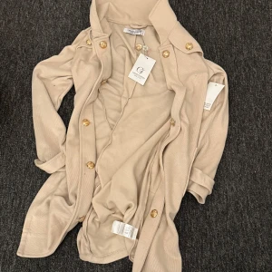 Beige kappa från Chiara Forni - Elegant beige kappa från Chiara Forni med gyllene knappar. Perfekt för en stilren look. Kappan har en klassisk design med långa ärmar och en mjuk känsla.