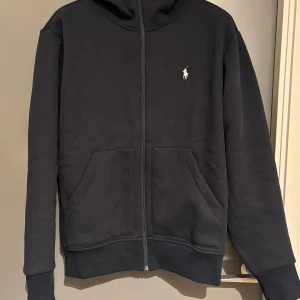 Svart hoodie från Ralph Lauren - Snygg svart hoodie från Ralph Lauren med dragkedja och klassisk logga på bröstet. Perfekt för en avslappnad stil med praktiska fickor framtill. Passar bra till både jeans och träningsbyxor. Skick är helt ny då den tyvärr satt för stort på mig.