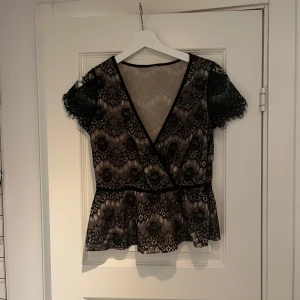 spetsblus med omlott  - Snygg svart spetsblus med omlott-design och korta ärmar. Perfekt för en kväll ute eller en fest. Blusen har ett vackert mönster och en feminin passform som framhäver midjan. Passar bra med jeans eller kjol för en chic look.