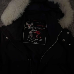 Svart jacka med pälsdetaljer från Moose Knuckles - Snygg svart jacka från Moose Knuckles med vit päls på huvan. Jackan har dragkedja och knappar framtill samt dekorativa detaljer på ärmarna. Perfekt för kalla dagar.