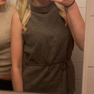 Brun ärmlös topp med knytband - Säljer en snygg brun ärmlös topp med hög krage och knytband i midjan. Perfekt för både vardag och fest! Toppen är i ett mjukt material. Köpte den här på plick, jag har använt den men den är fortfarande k väldigt bra skick. Hör av dig vid intresse!