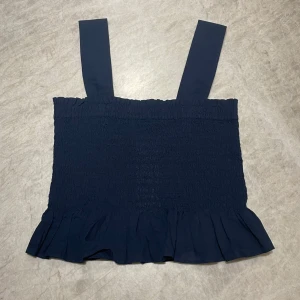 Mörkblå topp med volang - Säljer en superfin mörkblå topp med breda axelband och volangdetalj längst ner. Toppen är elastisk och sitter bekvämt på kroppen. Säljer den då den aldrig kommit till användning, prislappen sitter kvar.