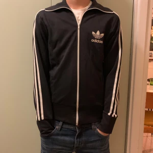 Svart träningsjacka från Adidas - Säljer en klassisk svart träningsjacka från Adidas med vita ränder längs ärmarna och logga på bröstet. Jackan har dragkedja. Den är i bra skick och passar till många olika outfits. 