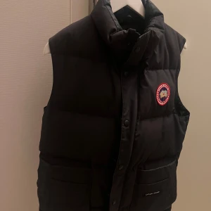 Svart dunväst från Canada Goose - Säljer en svart dunväst från Canada Goose med en broderad logotyp på bröstet. Västen har knappar och praktiska fickor framtill. Perfekt för kyligare dagar.
