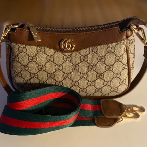 Beige och brun axelväska  - Snygg axelväska i beige och brunt. Väskan har en justerbar axelrem i grönt och rött, samt detaljer i guld. 