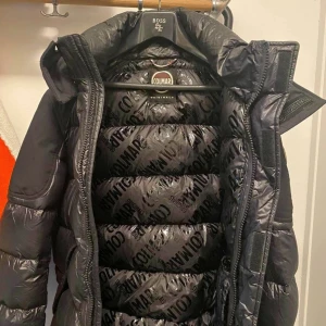 Svart dunjacka från Colmar - Snygg svart dunjacka från Colmar med hög krage och dragkedja. Jackan har en quiltad design och en logotyp på ärmen. Perfekt för kyliga dagar. Storlek M