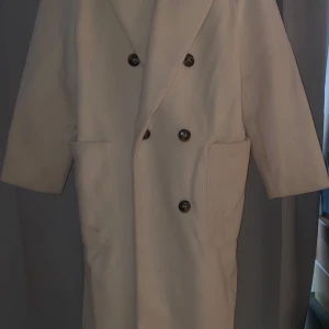 Beige kappa med knappar - Elegant beige kappa med dubbelknäppning och stora knappar. Den har en klassisk design med långa ärmar och två fickor framtill. Perfekt för kyligare dagar.