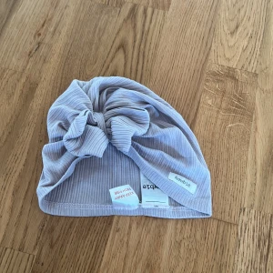 Lila ribbad mössa med rosett från Newbie - Söt lila ribbad mössa från Newbie med en stor rosett framtill. Perfekt för små barn i åldern 9-24 månader. Mysig och stilren design som passar till många tillfällen.