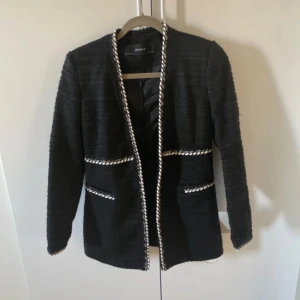 Svart kavaj med vita detaljer från Zara - Elegant svart kavaj från Zara med vita kantdetaljer. Den har en klassisk design med långärmade ärmar och två fickor framtill. Perfekt för en stilren look.