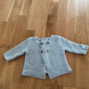 Grå stickad babykofta från Newbie - Söt grå stickad kofta för baby från Newbie. Den har dubbla knapprader framtill och långa ärmar. Perfekt för att hålla din lilla varm och mysig. 🐣
