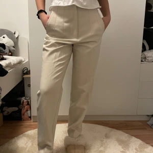 Beige kostymbyxor - Säljer ett par eleganta beige kostymbyxor i mycket bra skick. De är högmidjade med en klassisk straight passform som passar perfekt för både kontoret och festliga tillfällen. Byxorna har en stilren design med pressveck framtill och är tillverkade i ett bekvämt material. säljer för att dom är för stora 