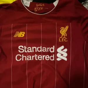 Röd Liverpool fotbollströja - Säljer en röd Liverpool fotbollströja från New Balance med korta ärmar. Den har vita ränder och tryck med 'Standard Chartered' på framsidan. På baksidan står det 'Mané' och nummer 10. Perfekt för fans av laget! 🏆