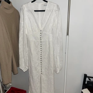Vit långklänning från Adoore - Elegant vit långklänning från Adoore med v-ringning och knappar framtill. Klänningen har långa ärmar och ett subtilt blommigt mönster. Perfekt för sommarens festligheter eller en stilfull vårdag.