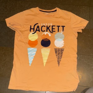 Orange t-shirt från Hackett London - Säljer en cool orange t-shirt från Hackett London med ett färgglatt glassmotiv på framsidan. Perfekt för sommarens äventyr! T-shirten är i bomull och har en normal passform med korta ärmar. Passar perfekt för både lek och vardag.