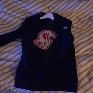 Mörkblå tröja från Moncler - Snygg mörkblå tröja från Moncler med ett stort rött och vitt logotyptryck på framsidan. Tröjan har långa ärmar och en rund halsringning. Perfekt för kyligare dagar.