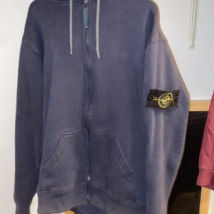 Mörkblå hoodie från Stone Island - Säljer en mörkblå hoodie från Stone Island med dragkedja och huva. Den har en broderad logga på ärmen och praktiska fickor framtill. Perfekt för en avslappnad stil. Kom gärna med frågor. Finns artikel kod