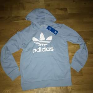 Säljer en ljusblå hoodie från Adidas med det klassiska trefoil-logotypen i vitt på framsidan. Tröjan har en bekväm passform med långa ärmar och en praktisk huva. Perfekt för en avslappnad stil.