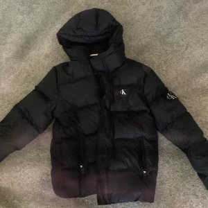 Svart pufferjacka från Calvin Klein Jeans - Snygg svart pufferjacka från Calvin Klein Jeans med huva och dragkedja. Jackan har märkets logga på bröstet och ärmen, vilket ger en stilren look. Perfekt för kyligare dagar.