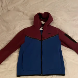 Tvåfärgad jacka från Nike - Snygg jacka från Nike i en cool tvåfärgad design med vinröd och blå. Jackan har en dragkedja framtill och en praktisk huva. Perfekt för en sportig look.