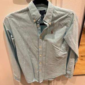 Ljusblå skjorta från Ralph Lauren - Snygg ljusblå skjorta från Ralph Lauren i slim fit. Skjortan har långa ärmar och knappar framtill. Perfekt för en stilren look med den ikoniska loggan på bröstet.