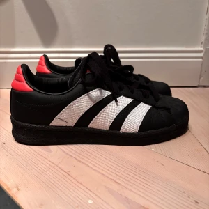 Adidas Wmns Superstar - Snygga svarta Adidas sneakers med klassiska vita ränder och en rosa detalj på hälen. Säker för att de är för små och kunde inte lämna tillbaka💗