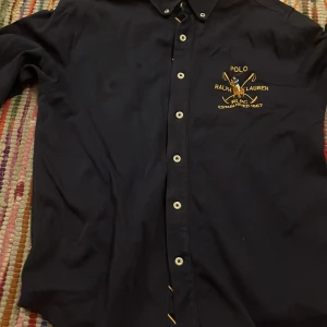 Svart skjorta från Ralph Lauren - Snygg svart skjorta från Ralph Lauren med broderad logga i gult på bröstet. Skjortan har knappar hela vägen och en klassisk krage. Perfekt för en stilren look.
