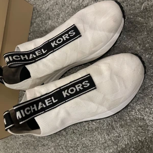 Vita sneakers från Michael Kors - Snygga vita sneakers från Michael Kors med elastisk passform och svart logotypband över foten. Inte trasiga någonstans och extremt bra skick. Använda ett få tal gånger