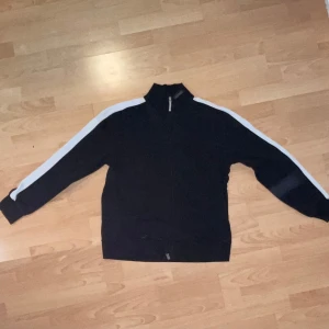 Svart jacka med vita ränder från Boss - Säljer en svart sweatshirt från Boss i herrstorlek L. Den har vita ränder längs ärmarna och en praktisk dragkedja framtill. 