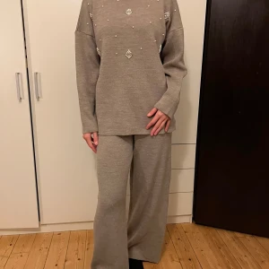 Beige stickad set med pärlor - Säljer en superfin beige stickad set med pärldetaljer. Tröjan är långärmad och har en avslappnad passform, perfekt för höst och vinter. Tröjan är både stilren och bekväm, byxorna är vida och långa. 