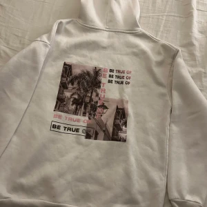 Vit hoodie med tryck - Säljer en vit hoodie med ett coolt tryck på baksidan. Trycket har en bild av palmer och texten 'BE TRUE'. Perfekt för en avslappnad stil och passar till alla årstider. Den har en klassisk känguruficka och en bekväm huva. Ganska tunn hoodie så inte så himla varm, passar nog bäst på sommaren när man är lite kylig💕