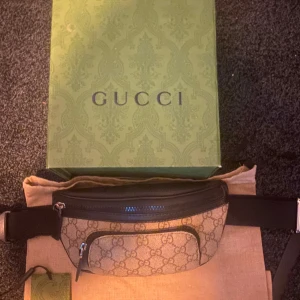 Mönstrad magväska från Gucci - Snygg magväska från Gucci med klassiskt mönster och svarta detaljer. Väskan har en dragkedja framtill och justerbart bälte för perfekt passform. Perfekt för att bära dina nödvändigheter med stil.