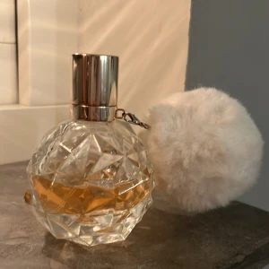 Ariana grande parfym (ARI) - Ariana grande parfym (ARI). Elegant parfymflaska med en unik, facetterad design och en rosé färgad kork. Flaskan har en dekorativ vit fluffig boll fäst vid korken, vilket ger en lyxig känsla. Perfekt för den som vill ha en stilfull detalj på sminkbordet. 