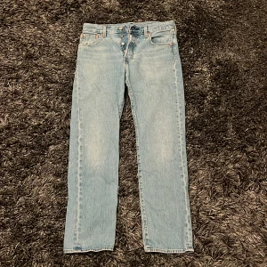 Ljusblå jeans från Levi's - Säljer ett par klassiska ljusblå Levi's jeans med knappgylf och raka ben. De har den ikoniska läderpatchen baktill och femficksdesign. Perfekta för en avslappnad stil.