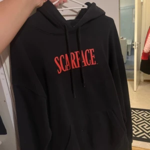 Svart Scarface hoodie från H&M - Cool svart hoodie från H&M med Scarface-tryck i rött på framsidan och ett stort motiv på baksidan. Perfekt för filmälskare som vill ha en bekväm och stilren look. Hoodien har en oversized passform och en klassisk känguruficka.