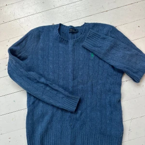 Blå kabelstickad tröja från Polo Ralph Lauren - Säljer en snygg blå kabelstickad tröja från Ralph Lauren. Tröjan är tillverkad av 90% merinoull och 10% kashmir, vilket gör den både mjuk och varm. Den har en klassisk rund hals och ett broderat grönt logotypmärke på bröstet.