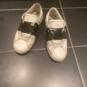 Valentino Garavani sneakers - Snygga Valentino Garavani sneakers i vitt med svarta detaljer. Skorna har en unik design med breda remmar över vristen och klassisk snörning. Perfekta för en stilren look. Storlek 42