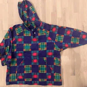 Vintage Crazy Fleece - Fleecen har halvlånga ärmar och har inga etiketter.   Säljer en färgglad hoodie med rutigt mönster i blått, rött och grönt. Tröjan har en stor huva och en avslappnad passform, perfekt för en chill dag. Den har en praktisk ficka framtill och knappar vid halsen.