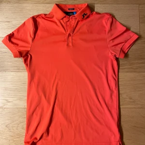 Orange  J.Lindeberg-polotröja med svarta detaljer - Tröjan har även en J.Lindeberg-logotyp på baksidan. Har används som golfpiké men är även snygg som vardags piké. Passar vanlig S eller tajt på en med storlek M. Nypris ca 900kr. Bara använd ett fåtal gånger så vara är nyskick!