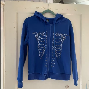 Blå hoodie med strassdetaljer från Shein - Säljer en cool blå hoodie från Shein med strassdetaljer som formar ett skelettmönster på framsidan. Den har en dragkedja och långa ärmar, perfekt för en avslappnad stil. Passar bra till höst och vår!