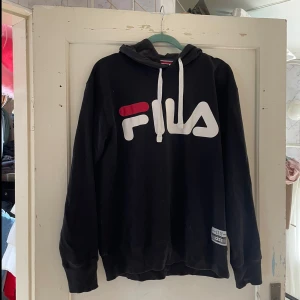 Svart hoodie från Fila - Säljer en svart hoodie från Fila i storlek M. Den har en stor Fila-logga i vitt och rött på framsidan och en klassisk känguruficka. Huvan har vita dragsnören och tröjan är perfekt för både höst och vinter. Superbekväm och stilren!