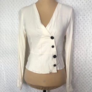 Vit omlottblus med knappar - Säljer en stilren vit omlottblus med snygga svarta knappar. Blusen har en v-ringad design och långa ärmar, perfekt för både vardag och fest. Materialet känns mjukt och bekvämt mot huden. Passar bra till både jeans och kjol!