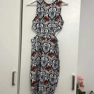 Mönstrad klänning från Amis - Säljer en superfin klänning från Amisu med ett färgglatt blommönster i rött, blått och gult på en vit bakgrund. Klänningen är ärmlös och har en smickrande passform med en dragkedja i ryggen. Perfekt för sommarens fester eller en dag på stan!